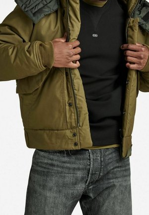 Homme portant une veste matelassée vert olive par-dessus un sweat-shirt noir et un jean gris délavé, ajustant la fermeture éclair de la veste.