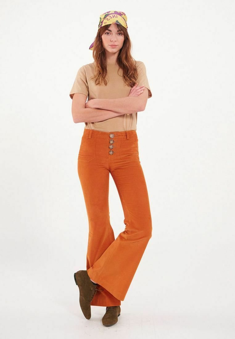 T-shirt tan associé à un pantalon en velours côtelé orange évasé avec fermeture à cinq boutons, mannequin se tenant debout portant un foulard à motifs et des chaussures marron.