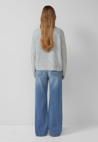 Vrouw met lang golvend haar, gekleed in een lichtgrijze gebreide trui en wijde vervaagde blauwe spijkerbroek, staand tegen een effen lichte muur.