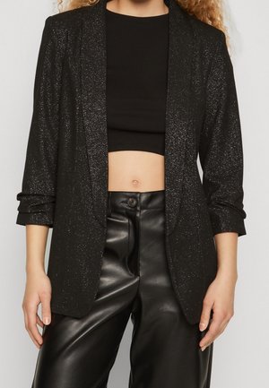 Mujer vistiendo un blazer negro con brillo, top negro recortado y pantalones de cuero negros de talle alto, con las manos relajadas a los costados.