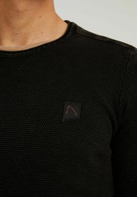 Czarny, teksturowany, długi rękaw top z okrągłym dekoltem oraz małą czarną łatką z logiem, na której znajduje się pomarańczowy wzór na klatce piersiowej.