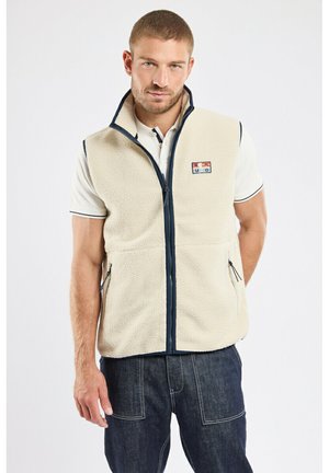 Homme portant un gilet en polaire crème sur un polo blanc et un jean bleu foncé, debout devant un fond clair uni.