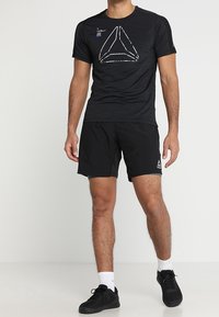 Black athletic t-shirt with a geometric logo design in white, paired with black shorts and black shoes. Textured fabric, short sleeves.