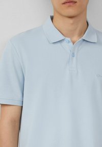 Helles blaues Poloshirt aus strukturiertem Material, mit Kragen, zwei Knöpfen und einem kleinen Logo auf der linken Seite.