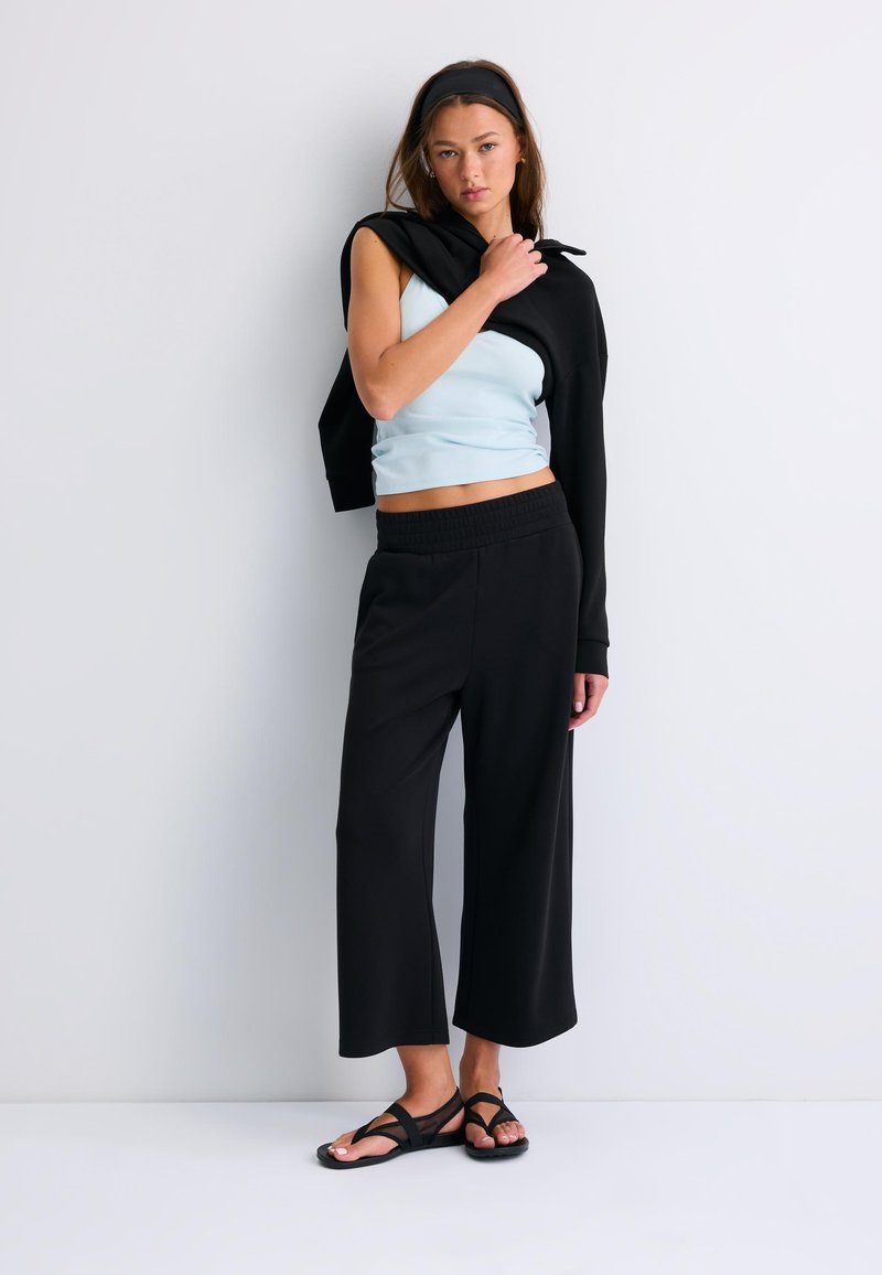 Jeune femme appuyée contre un mur blanc, portant un haut sans manches bleu clair, un pantalon large noir, des sandales noires et un pull noir sur l'épaule.
