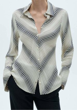 Femme portant une chemise boutonnée rayée noir et blanc avec des lignes diagonales, associée à un pantalon noir, mains dans les poches.