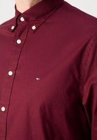 Bordowa koszula bawełniana z kołnierzykiem zapinanym na guziki, z białymi guzikami i małym logo w kolorach czerwonym, białym i niebieskim na piersi.