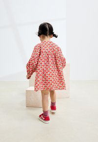 Blomstret kjole med røde, grønne og pink mønstre, ballonærmer, kravedetaljer, kombineret med røde sneakers og pink sokker på en lys overflade.