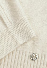 Tissu tricoté couleur crème avec des poignets côtelés et une petite épingle ronde en argent avec un design de cœur stylisé.