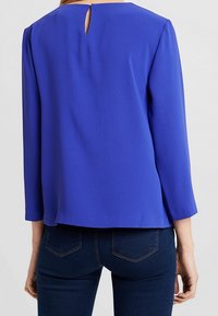 Maglietta blu a maniche lunghe con scollatura tonda e apertura a chiave sul retro. Tessuto liscio, vestibilità leggermente ampia e dettagli cuciti.