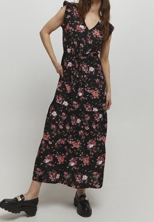 Vrouw draagt een zwarte mouwloze maxijurk met roze en witte bloemenprint en zwarte grove loafers, staand tegen een effen achtergrond.