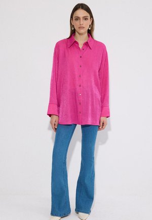 Žena stojaca, oblečená v jasne ružovej košeli s dlhými rukávmi a modrých rozšírených džínsoch, na jednoduchom bielom pozadí.