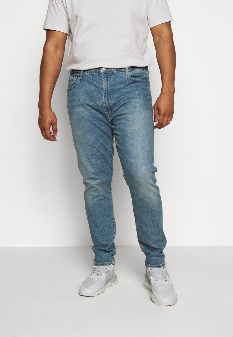 Light blue denim jeans with a slim fit, faded finish, and five pockets, worn with a white t-shirt and gray athletic sneakers.