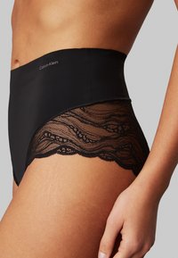 Korkean vyötärön mustat alushousut, joissa on sileä, yhtenäinen kangas ja monimutkaista pitsiä reunoilla. Ominaisuutena hienovarainen Calvin Klein -logointi.