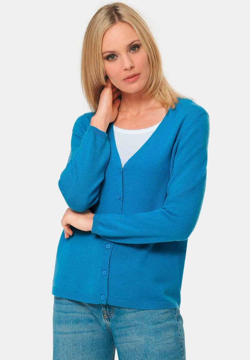 Türkise Strickjacke mit V-Ausschnitt, langen Ärmeln und vorderer Knopfverschluss. Weicher Stoff und gerippte Bündchen, kombiniert mit einem weißen Oberteil darunter.