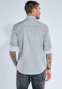 Man with tattoos on both forearms wearing a light grey patterned long-sleeve shirt with sleeves rolled up and dark jeans, facing away.