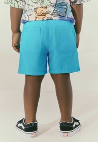 Pantaloni corti turchesi realizzati in tessuto leggero, dotati di due tasche posteriori, abbinati a sneakers Vans nere e bianche.