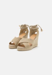 Sandales compensées beiges avec des brides tressées croisées, dessus texturé et liens à la cheville ornés de franges. Semelle en fibres naturelles avec une légère élévation.