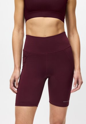 Vrouw draagt donker bordeauxrode atletiekshorts met hoge taille en bijpassende sportbeha, staande met ontspannen armen langs het lichaam.