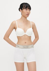 Bijela grudnjak s dubokim izrezom i tankim naramenicama u kombinaciji s bijelim boksericama koje imaju crni i bež Calvin Klein logo na elastičnom pojasu.