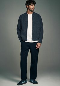 Donkergrijze bomberjack met een rits aan de voorkant, gecombineerd met een wit T-shirt, marineblauwe broek en marineblauwe sneakers met blauwe strepen.