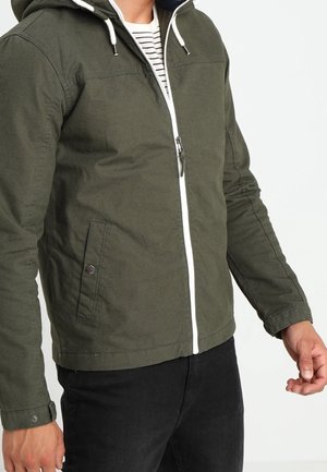 Homme portant une veste zippée vert olive avec des cordons blancs sur une chemise rayée, associée à un pantalon noir, visible des épaules aux hanches.