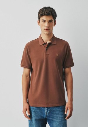 Junger Mann mit kurzen lockigen Haaren, der ein braunes Poloshirt und blaue Jeans trägt und vor einem schlichten hellen Hintergrund steht.