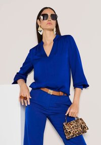 Königsblaue Bluse mit V-Ausschnitt und Dreiviertelärmeln; kombiniert mit passenden Hosen und einem braunen Gürtel. Hält eine Handtasche mit Leopardenmuster.
