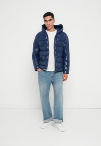 Jachetă puffer bleumarin cu finisaj lucios, închidere cu fermoar și glugă. Modelul poartă un tricou alb, blugi deschiși la culoare și adidași albi.