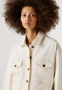 Šviesiai kreminė džinsinė striukė su apykakle ir sagomis, su dviem krūtinės kišenėmis, kontrastingais siūlais ir tekstūruota medžiaga.