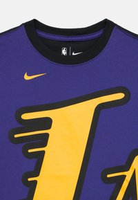 Μωβ βαμβακερό T-shirt με μεγάλο μαύρο και κίτρινο γράμμα "L", με μαύρο λαιμόκοψη και μικρό λογότυπο Nike στο λαιμό.