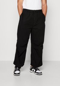 Schwarze, locker sitzende Cargo-Hose mit elastischem Bund, Seitentaschen und glatter Textur, kombiniert mit schwarzen und weißen Sneakers.