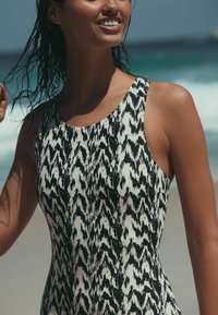 Moteris su šlapiais plaukais, dėvinti juodai baltos spalvų raštuotą rankovių neturinčią maudymosi kostiumėlį saulėtoje paplūdimyje.