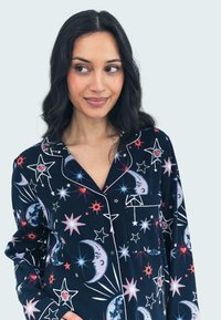 Marina pyjamasset har ett mönster av stjärnor och månar i vitt, rött och blått, med en slagkrage och en ficka på vänster bröst. Mjuk tygtextur.