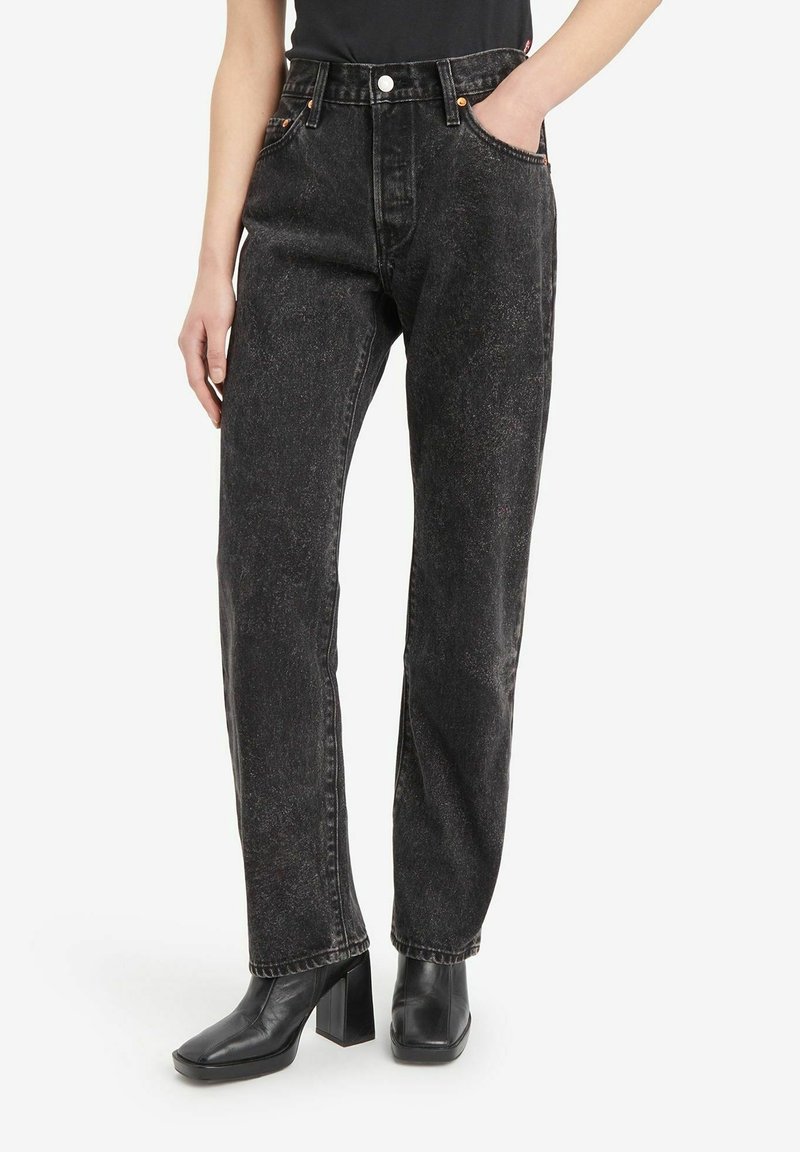 Personne portant un jean noir taille haute à jambes droites avec bouton et fermeture éclair, associé à des bottes noires à talons et bout carré.