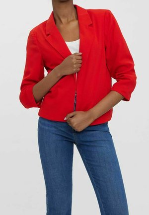 Frau trägt einen knallroten Blazer über einem weißen Top und blauen Jeans, hält den Blazer mit beiden Händen geschlossen.