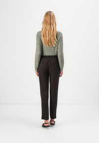 Pantalon marron à taille élastique associé à un pull vert texturé, vu de dos. Cheveux longs et ondulés. Modèle de chaussure simple visible.