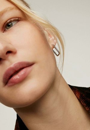 Pendiente de aro lineal dorado con una ligera curva, sujeto a la oreja de un modelo, mostrado sobre un fondo neutro con una iluminación suave.