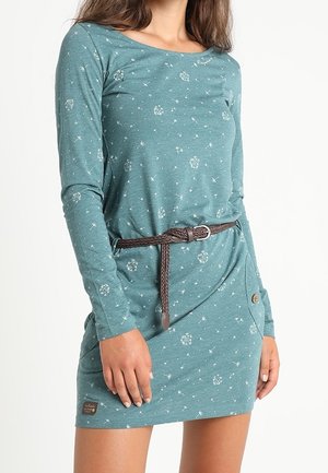 Donna con un abito verde teal a maniche lunghe con stampa a soffione, cintura intrecciata marrone, e tasca laterale con bottone, in piedi su uno sfondo bianco.