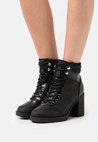 Botines negros con parte superior texturizada, acabado brillante, diseño de cordones, ribete de pelo sintético y tacón cuadrado grueso.