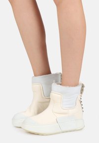 Vita läderankelboots med en texturerad vit krage, som har silvernitar och en gummisula. Den texturerade ytan tillför visuellt intresse.