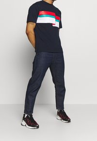 Marinblå T-shirt med ett frampaneldesign som har röda, vita och turkosa ränder; ihop med mörk denimjeans och svarta sneakers.