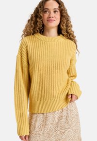 Pull en tricot jaune avec une texture côtelée, encolure ronde et manches larges, associé à une jupe beige à motifs floraux.