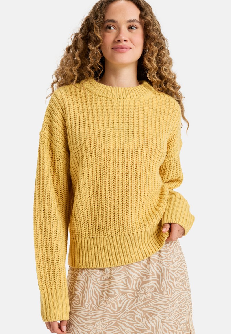 Pull en tricot jaune avec une texture côtelée, encolure ronde et manches larges, associé à une jupe beige à motifs floraux.