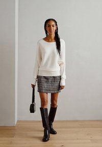 Pull blanc à côtes, jupe mini à motifs noir et blanc, bottes noires hautes. Le mannequin tient un petit sac à main noir, debout sur un sol en bois.