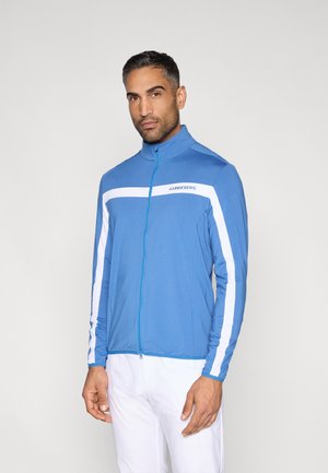 Modrá bunda so zipsom, vybavená bielymi pruhmi, dlhými rukávmi, stojatým golierom a ľahkým materiálom. Názov značky "J. Lindeberg" je viditeľný na hrudi.