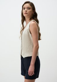Gilet en lin beige clair avec un décolleté en V, sans manches, fermeture à boutons et coupe ajustée, associé à un short bleu marine.