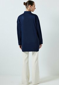 Cămașă albastru închis, cu mâneci lungi, cu guler, cu o croială lejeră și textură moale. Asortată cu pantaloni cremoși, evazați.