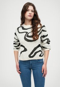 Baltas megztinis su juodais abstrakčiais raštais, apvaliu iškirpimu ir tris ketvirčius ilgomis rankovėmis, derinamas su mėlynais džinsais. Tekstūruota medžiaga.