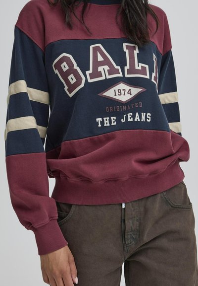 Karmiinpunane ja tumesinine sviiter valge kirjatüübiga, millel on triibud varrukatel ja soonikhems. Kujundus sisaldab sõna "BALI" ja aastat "1974."